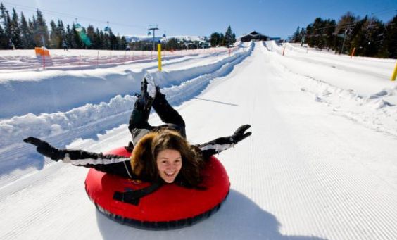 Eine Person lächelt, während sie an einem klaren Wintertag auf einem roten Schneeschlauch mit ausgestreckten Armen einen verschneiten Hügel hinunterrutscht.