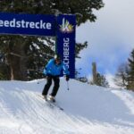 Ein Skifahrer in einer blauen Jacke fährt eine verschneite Piste unter einem Schild mit der Aufschrift "Speedstrecke Kreischberg" hinunter, umgeben von Bäumen.