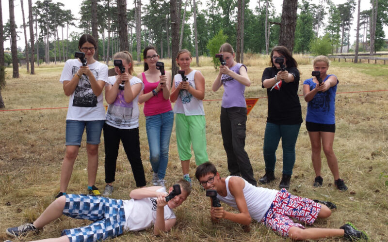 Eine Gruppe von neun jungen Leuten posiert im Freien mit Spielzeugpistolen; sieben stehen, während zwei vor ihnen auf dem Boden liegen. Im Hintergrund sind Bäume und trockenes Gras zu sehen.
