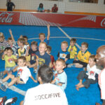 Eine Gruppe von Kindern sitzt auf einem blauen Hallenfußballfeld, lächelt und jubelt, während zwei Erwachsene in "SOCCAFIVE"-Shirts daneben knien.