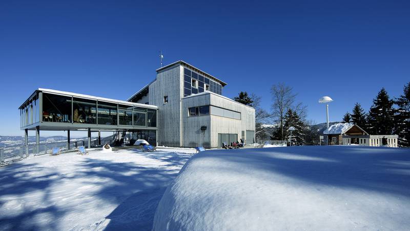 Modernes Gebäude mit großen Fenstern, umgeben von Schnee, Bäumen und einem klaren blauen Himmel, mit einem kleinen Gebäude im Hintergrund.
