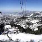 Eine rote Seilbahn fährt über eine verschneite Berglandschaft mit verstreuten Häusern und einer in der Ferne sichtbaren Stadt unter einem klaren blauen Himmel.