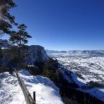 Schneebedeckter Aussichtspunkt auf einen Berg mit Bäumen auf der linken Seite, einem Holzgeländer und einem breiten Tal unter einem klaren blauen Himmel mit hellem Sonnenlicht.