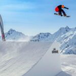 Ein Snowboarder führt in der Luft einen Sprung von einer Schneerampe mit schneebedeckten Bergen im Hintergrund aus.