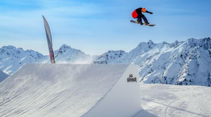 Ein Snowboarder führt in der Luft einen Sprung von einer Schneerampe mit schneebedeckten Bergen im Hintergrund aus.