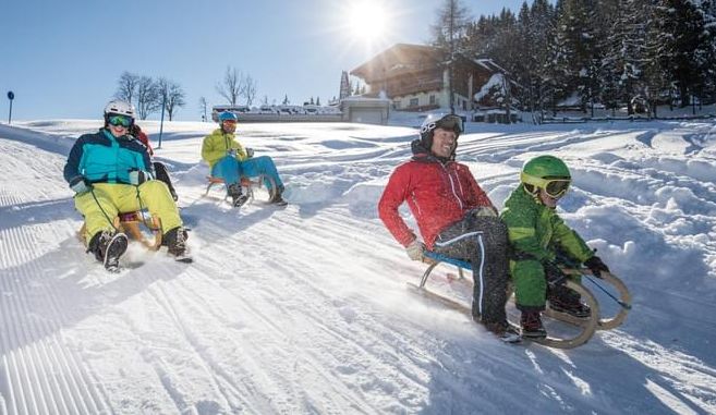 Vier Personen rodeln auf einer verschneiten Piste im hellen Wintersonnenlicht bergab, mit einem Holzhaus und Bäumen im Hintergrund.