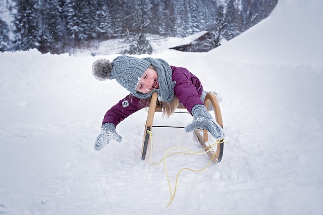 Eine Person in Winterkleidung fährt auf einem Holzschlitten einen verschneiten Hügel hinunter, wobei sie sich mit behandschuhten Händen nach vorne streckt. Im Hintergrund sind schneebedeckte Bäume zu sehen.