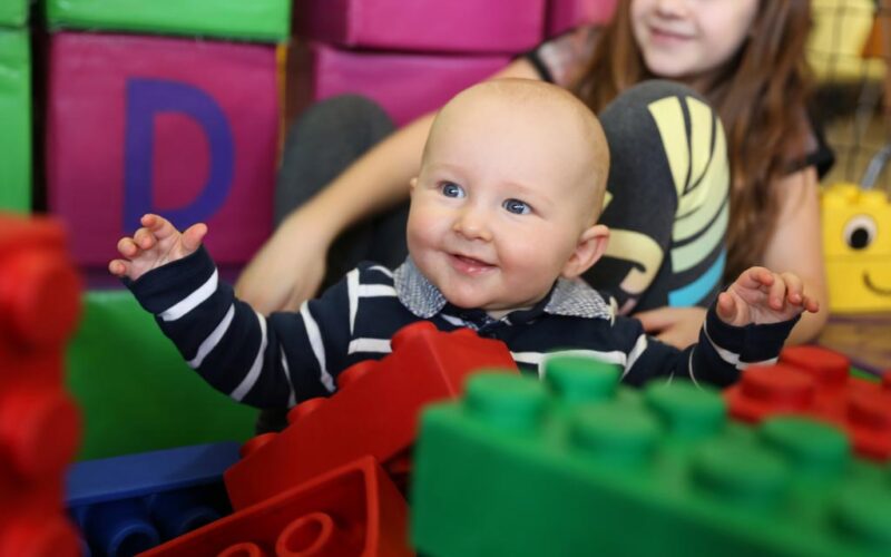 Ein Baby in einem gestreiften Hemd sitzt lächelnd zwischen großen bunten Spielzeugblöcken, im Hintergrund ein älteres Kind.