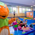 Eine Person in einem orangefarbenen Eichhörnchen-Maskottchenkostüm steht in der Nähe von Kindern und Erwachsenen, die mit großen Blöcken und Spielzeug in einem Indoor-Spielbereich spielen.