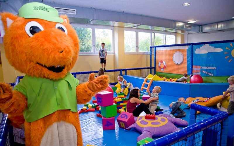 Eine Person in einem orangefarbenen Eichhörnchen-Maskottchenkostüm steht in der Nähe von Kindern und Erwachsenen, die mit großen Blöcken und Spielzeug in einem Indoor-Spielbereich spielen.