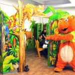 Eine Person in einem orangefarbenen Tierkostüm steht vor einem farbenfrohen, dschungelartigen Spielbereich, in dem Kinder an einem Tisch sitzen.