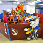 Kinder sitzen an einem Tisch in einem Spielbereich mit maritimer Dekoration, während ein Erwachsener und eine Person in einem orangefarbenen Katzenkostüm in der Nähe stehen.