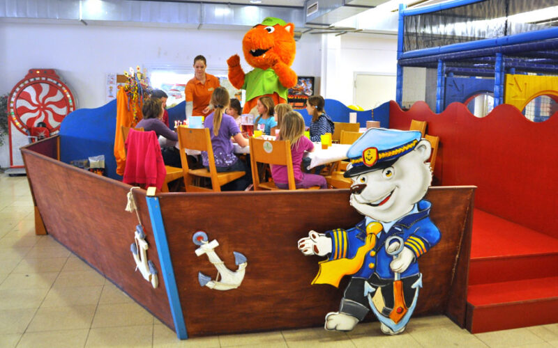 Kinder sitzen an einem Tisch in einem Spielbereich mit maritimer Dekoration, während ein Erwachsener und eine Person in einem orangefarbenen Katzenkostüm in der Nähe stehen.