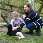 Eine Frau und ein Kind hocken neben einem schwarz-weißen Lamm und streicheln es in der Nähe eines Zauns auf einem Bauernhof im Freien.