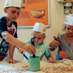 Drei Kinder mit Papierhüten bestreichen an einem Tisch Teig mit Eigelb und nehmen an einer Backaktivität teil.