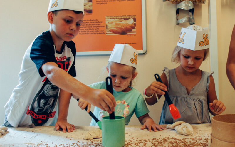 Drei Kinder mit Papierhüten bestreichen an einem Tisch Teig mit Eigelb und nehmen an einer Backaktivität teil.