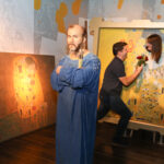 Eine Wachsfigur eines bärtigen Mannes steht vor Gemälden, während im Hintergrund ein Paar als Teil des Kunstwerks posiert.