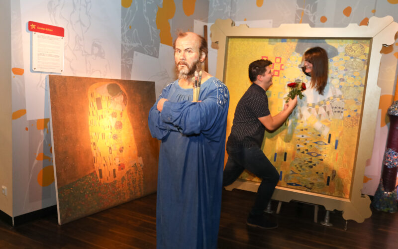 Eine Wachsfigur eines bärtigen Mannes steht vor Gemälden, während im Hintergrund ein Paar als Teil des Kunstwerks posiert.
