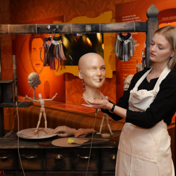 Eine Frau in einer Schürze hält einen Schaufensterpuppenkopf in einer Werkstatt, in der Schaufensterpuppenteile, Werkzeuge und Puppen auf einem Holztisch ausgestellt sind.
