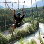 Zwei Personen mit Helmen und Klettergurten navigieren in einem Hochseilgarten über einem Fluss, mit Bäumen und Bergen im Hintergrund.