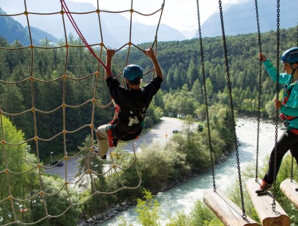 Zwei Personen mit Helmen und Klettergurten navigieren in einem Hochseilgarten über einem Fluss, mit Bäumen und Bergen im Hintergrund.