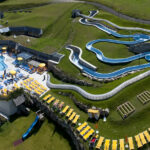 Luftaufnahme eines Wasserparks im Freien mit einem Poolbereich, Sonnenliegen und einer gewundenen blauen Wasserrutsche auf einem grasbewachsenen Hügel.