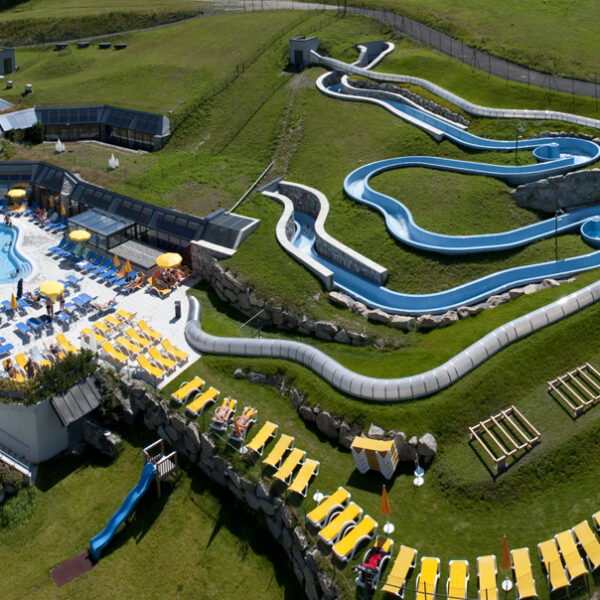 Luftaufnahme eines Wasserparks im Freien mit einem Poolbereich, Sonnenliegen und einer gewundenen blauen Wasserrutsche auf einem grasbewachsenen Hügel.