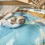 Hallenbad mit klarem Wasser, einem Felsen in der Mitte, Liegestühlen an den Seiten und zwei Personen, die auf einer kleinen Insel zusammensitzen.
