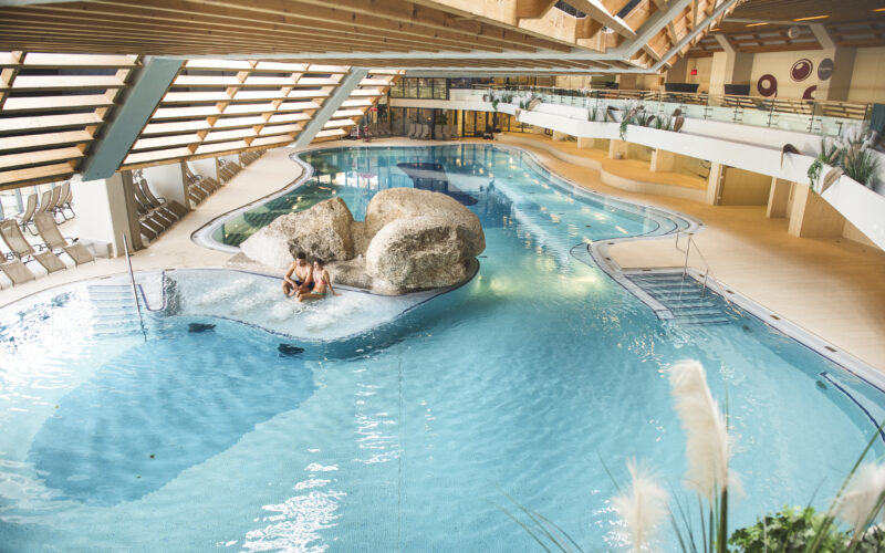Hallenbad mit klarem Wasser, einem Felsen in der Mitte, Liegestühlen an den Seiten und zwei Personen, die auf einer kleinen Insel zusammensitzen.