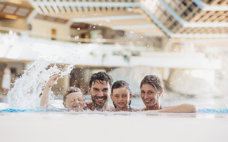 Ein Mann, eine Frau und zwei Kinder lächeln gemeinsam am Rande eines Hallenbades, während ein Kind mit Wasser spritzt.