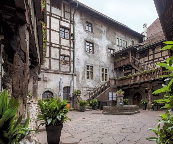 Ein historischer Steinhof mit Topfpflanzen, einem zentralen Brunnen, Holzbalkonen und Treppen, umgeben von Fachwerkwänden und Fenstern.