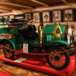 Ein grüner Oldtimer steht auf einer roten Plattform in einem Museum, umgeben von Informationsschildern, gerahmten Dokumenten und einem roten Absperrseil.