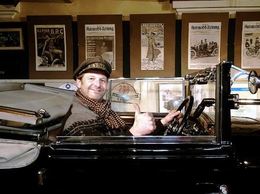 Ein Mann mit Mütze und Schal sitzt auf dem Fahrersitz eines Oldtimers, lächelt und zeigt den Daumen nach oben; an der Wand hinter ihm hängen alte Autoposter.