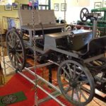 Im Innenraum ist ein Oldtimer aus Holz mit großen Speichenrädern ausgestellt, umgeben von Museumsdekor und gerahmten Fotos an den Wänden.