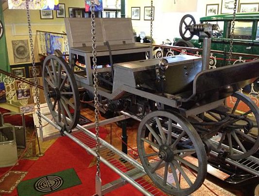 Im Innenraum ist ein Oldtimer aus Holz mit großen Speichenrädern ausgestellt, umgeben von Museumsdekor und gerahmten Fotos an den Wänden.
