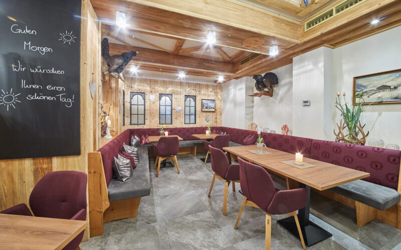 Ein gemütlicher Restaurantbereich mit kastanienbraunen, gepolsterten Sitzbänken, Holztischen, dekorierten Wänden und einer Kreidetafel, auf der ein deutscher Gruß steht.