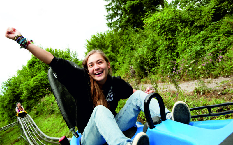 Eine junge Person fährt im Freien auf einem blauen Alpine Coaster, lächelt und hebt einen Arm, während im Hintergrund eine Grünfläche und ein anderer Fahrer zu sehen sind.
