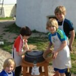 Vier Kinder in Schürzen stehen im Freien um eine Steinmühle herum. Ein Kind mahlt Getreide, andere schauen zu oder helfen mit. Im Hintergrund ist ein weißes Gebäude zu sehen.