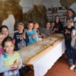 Eine Gruppe Kinder und ein Erwachsener stehen lächelnd um einen langen, mit Mehl bedeckten Holztisch herum und formen Teig in einem rustikalen Raum mit Steinwänden und Regalen.