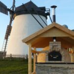 Im Hintergrund steht eine weiße Windmühle, im Vordergrund ein holzbefeuerter Außenofen unter einem Holzunterstand.