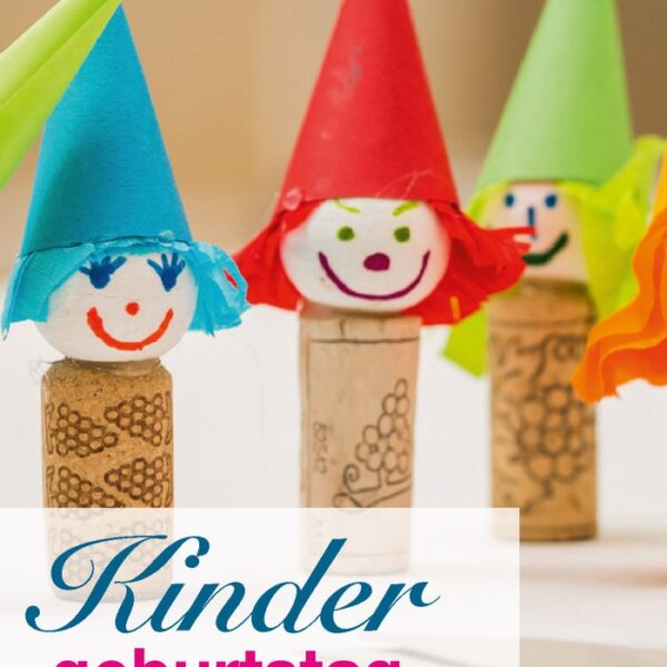 Drei Korkpuppen mit bunten Papierhüten und bemalten Gesichtern stehen aufrecht auf einem Tisch. Der deutsche Text lautet: „Kindergeburtstag im KunstTRAUMstift.“.