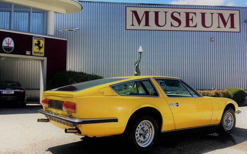 Ein gelber klassischer Sportwagen parkt vor einem Gebäude mit einem großen MUSEUM"-Schild und Automobillogos an der Wand.