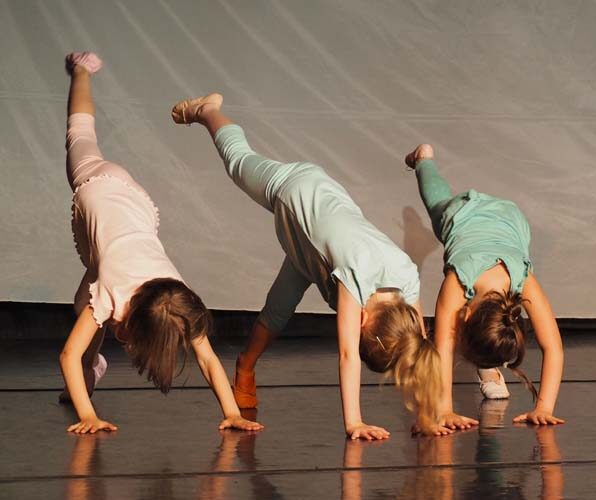 Drei kleine Kinder führen auf der Bühne eine Tanzeinlage auf, wobei jedes auf einem Bein und einer Hand balanciert und das andere Bein nach hinten angehoben ist.