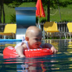 Ein Baby sitzt in einem roten aufblasbaren Ring, der in einem Pool schwimmt, umgeben von gelben Liegestühlen und orangefarbenen Sonnenschirmen an einem sonnigen Tag.