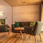 Ein modernes Hotelzimmer mit Holzfußboden, einem Bett, einem grünen Sofa, zwei grauen Sesseln um einen runden Tisch und großen Fenstern, die auf einen Balkon mit Blick auf den Wald führen.