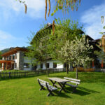Hinter einer eingezäunten Wiese mit Picknicktischen und blühenden Bäumen steht unter blauem Himmel ein Holzgebäude mit Schrägdach.