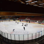 Mehrere Personen laufen Schlittschuh und spielen auf einer überdachten Eisbahn bei künstlicher Beleuchtung, die von leeren Tribünen umgeben ist.