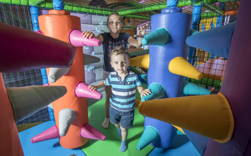 Zwei Kinder spielen auf einem Indoor-Spielplatz zwischen großen, gepolsterten Rollen mit bunten, weichen Stacheln in einem Spielbereich mit Netzen.