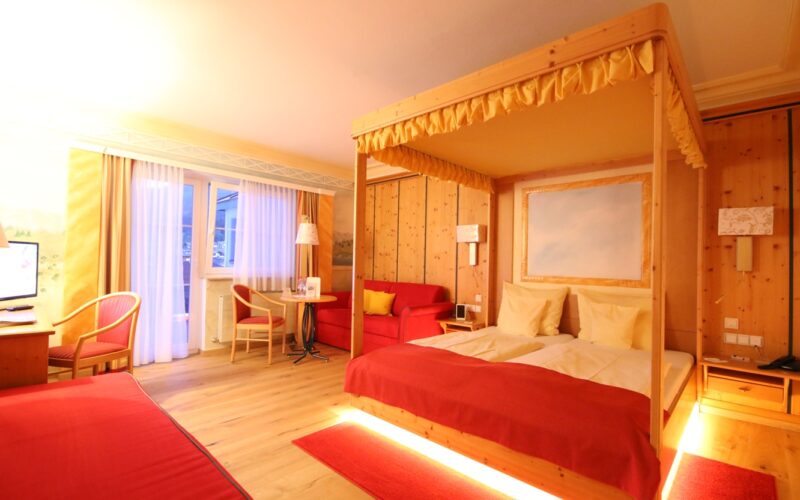 Ein Hotelzimmer mit Holzwänden, einem Himmelbett, rotem Bettzeug, einem Schreibtisch, einem Sofa, einem kleinen Tisch mit Stühlen und einer Balkontür, durch die Tageslicht einfällt.