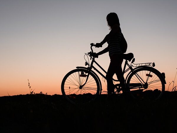 Silhouette einer Person, die bei Sonnenuntergang im Freien neben einem Fahrrad steht, mit einem orangefarbenen und lila Himmel im Hintergrund.